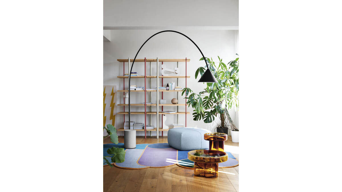 Lampada da Terra Ozz Floor nera con base in calcestruzzo in una stanza con tavolini orange soda, tappeto, libreria, pianta ed un pouf.