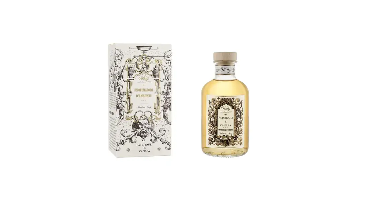 Una bottiglia in vetro di Patchouli E Canapa Profumatore d'ambiente, riempita di liquido giallo ambra, fornita di etichetta decorativa e tappo in legno. Accanto la sua confezione decorata da illustrazioni bianco, nero e oro.