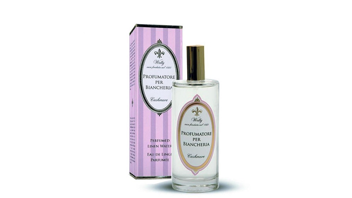 Una bottiglia in vetro di Profumatore per biancheria Cashmere riempita di liquido trasparente, fornita di etichetta decorativa. Accanto la sua confezione decorata da illustrazioni color viola, lillà e bianco.