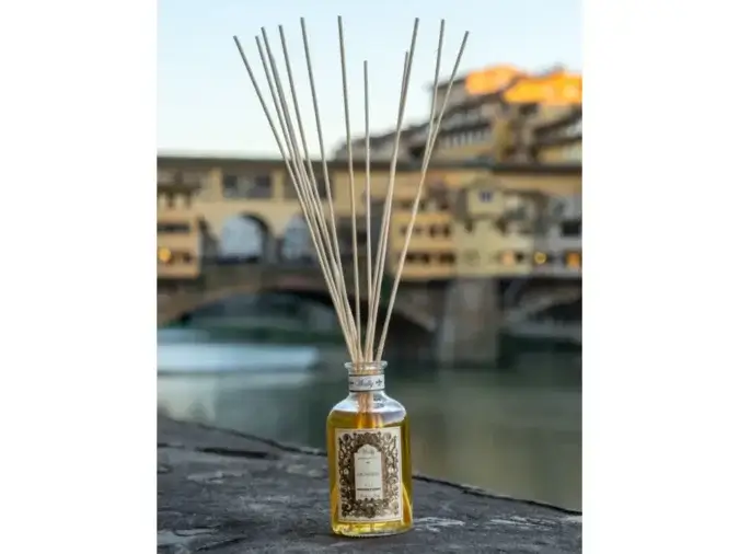 Una bottiglia in vetro di Signoria Profumatore d'ambiente, riempita di liquido giallo ambra con etichetta decorativa e bastoncini in legno. La bottiglia è posta su un muretto con Ponte Vecchio di Firenze sullo sfondo.
