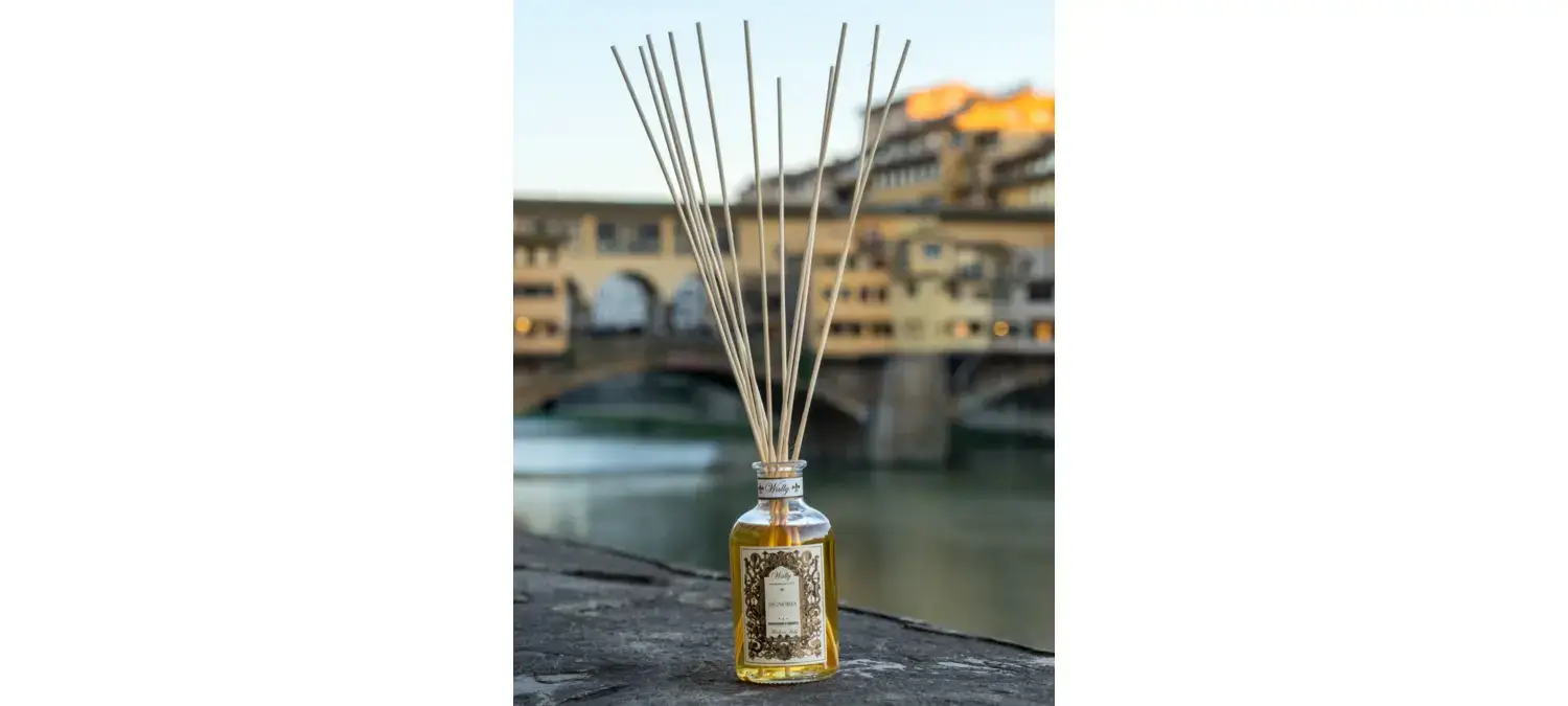 Una bottiglia in vetro di Signoria Profumatore d'ambiente, riempita di liquido giallo ambra con etichetta decorativa e bastoncini in legno. La bottiglia è posta su un muretto con Ponte Vecchio di Firenze sullo sfondo.