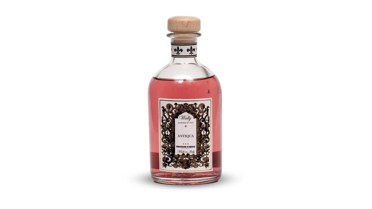 Una bottiglia in vetro di Antiqua Profumatore d'ambiente, riempita di liquido color rosa, fornita di etichetta decorativa e tappo in legno.