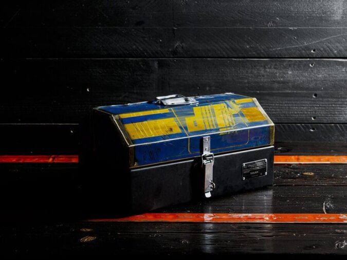Cassetta degli attrezzi GT Toolbox in lamiera di bidone appoggiata su un piano di legno nero con fasce arancioni.