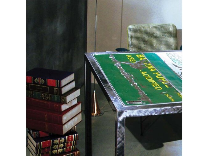 Leggero Tavolo fisso in lamiera verde di bidone con scritte gialle posto in living room con una sedie, dei libri ed una lampada.