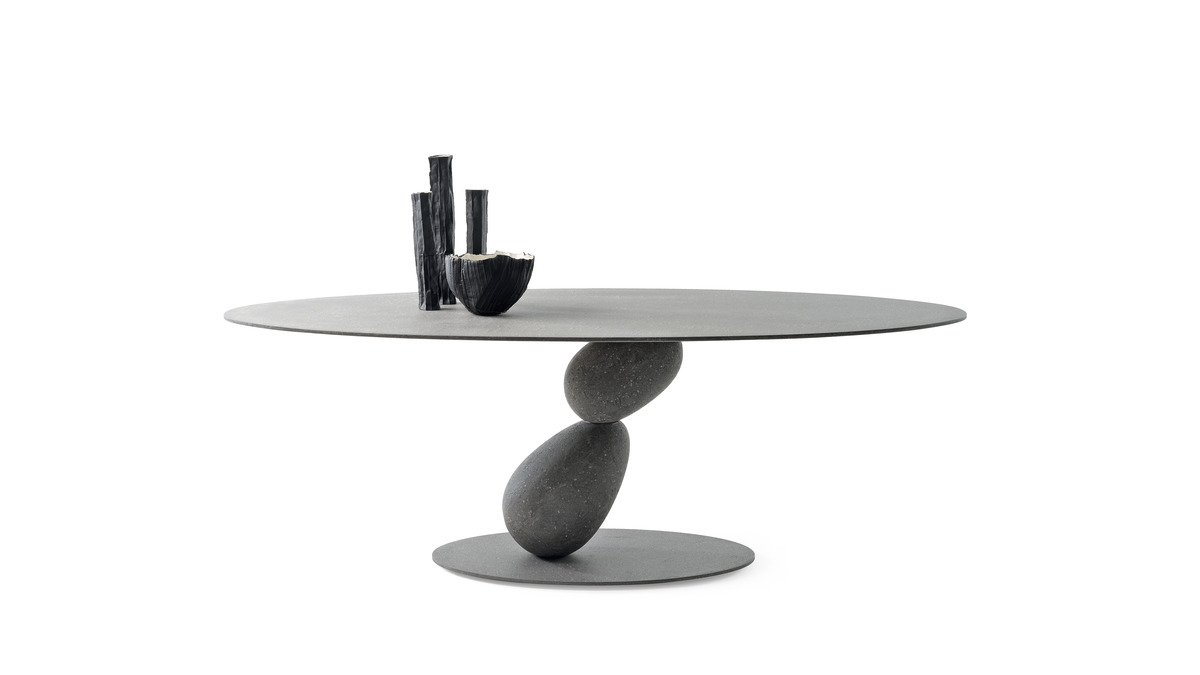 Tavolo Matera con piano circolare in finitura grigio urbano, bilanciato su una base scultorea composta da pietre lisce impilate. Vari vasi neri sono posti sul tavolo.