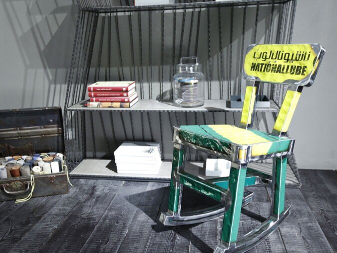 Sedia a dondolo Mecedora in lamiera di bidone verde e gialla con scritte nere, situata in una living room con una libreria in ferro, ornata da libri e contenitori.