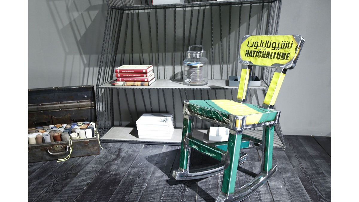 Sedia a dondolo Mecedora in lamiera di bidone verde e gialla con scritte nere, situata in una living room con una libreria in ferro, ornata da libri e contenitori.
