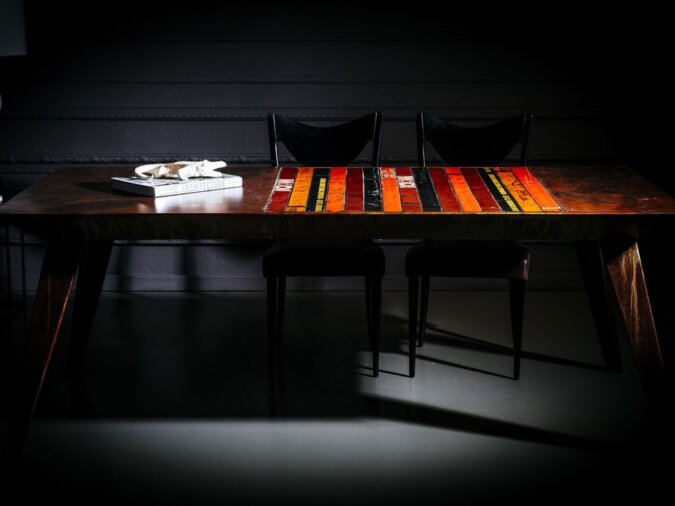 Tavolo Pitagora in lamiera di bidone in una dining room in penombra con due sedie nere ed un libro sul piano.