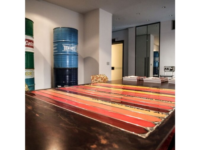 Primo piano del tavolo Pitagora in lamiera di bidone in una dining room con dei libri appoggiati sul ripiano, sedie in lamiera e bidoni di petrolio sullo sfondo.