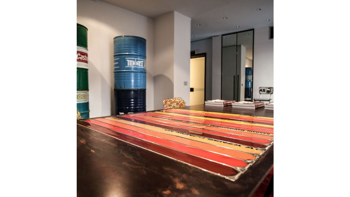 Primo piano del tavolo Pitagora in lamiera di bidone in una dining room con dei libri appoggiati sul ripiano, sedie in lamiera e bidoni di petrolio sullo sfondo.