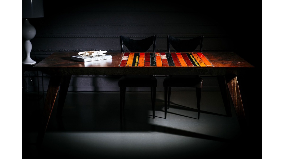Tavolo Pitagora in lamiera di bidone in una dining room in penombra con due sedie nere ed un libro sul piano.