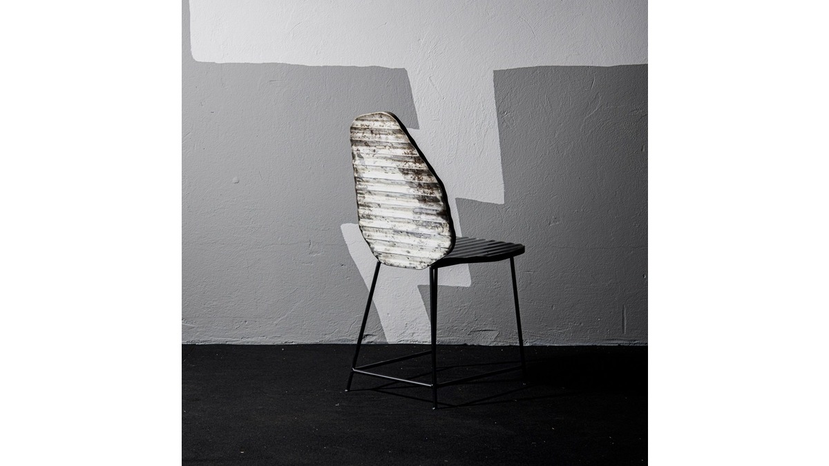 Vista posteriore della sedia Rock Chair in lamiera di bidone in una stanza con pavimento nero e muro in cemento grigio.