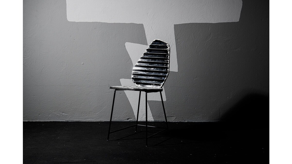 Vista Frontale della Sedia Rock Chair in lamiera di bidone in una stanza con pavimento nero e muro in cemento grigio.