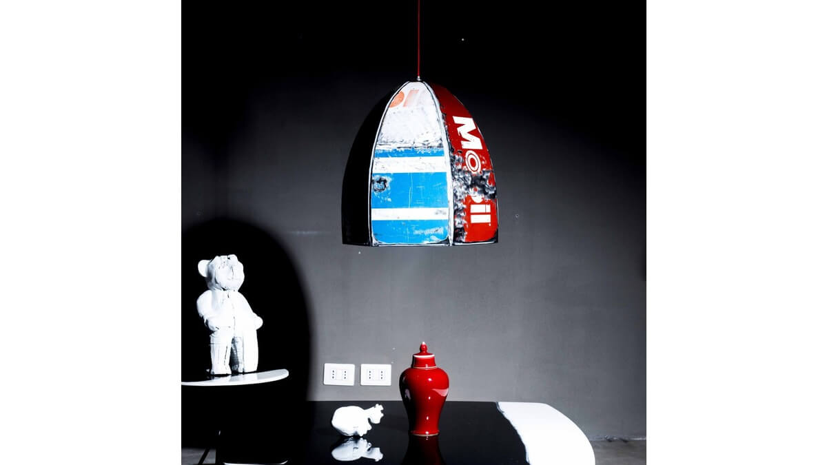 Pendant Lamp Rugby in lamiera di bidone blu e rosso con scritta Mobil in living room con un tavolo nero, sul quale sono poggiati un vaso rosso e altri oggetti, di cui un orso in porcellana.