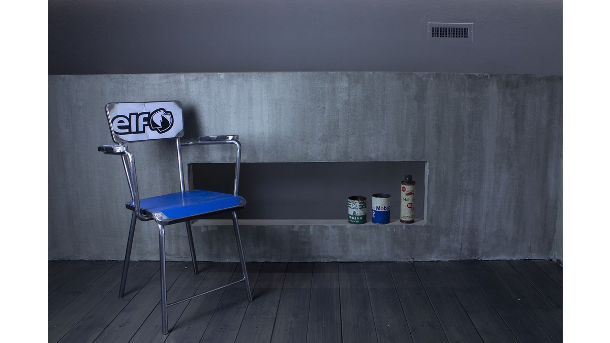 Sedia Secca con braccioli in lamiera di bidone con seduta blu e schienale bianco con scritta "ELF" di fronte a un muro di cemento grigio con una nicchia incassata, che ospita vari contenitori di latta.