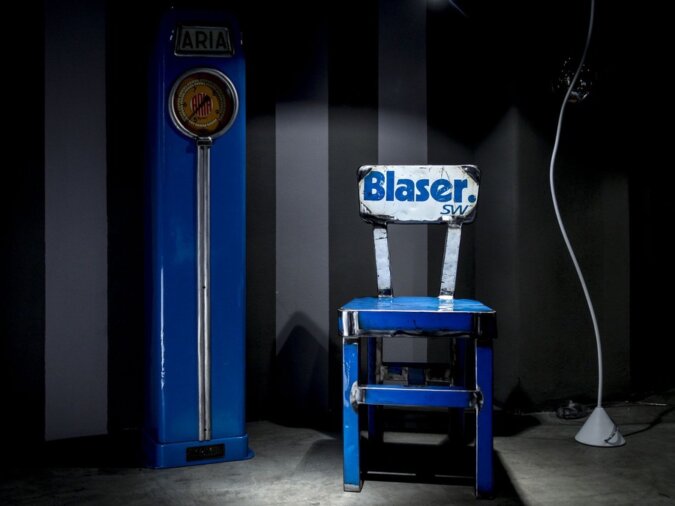 Sedia dora in lamiera di bidone blu con scritta Blaser in una stanza poco illuminata, affiancata da una pompa di benzina aria vintage e una lampada da terra.