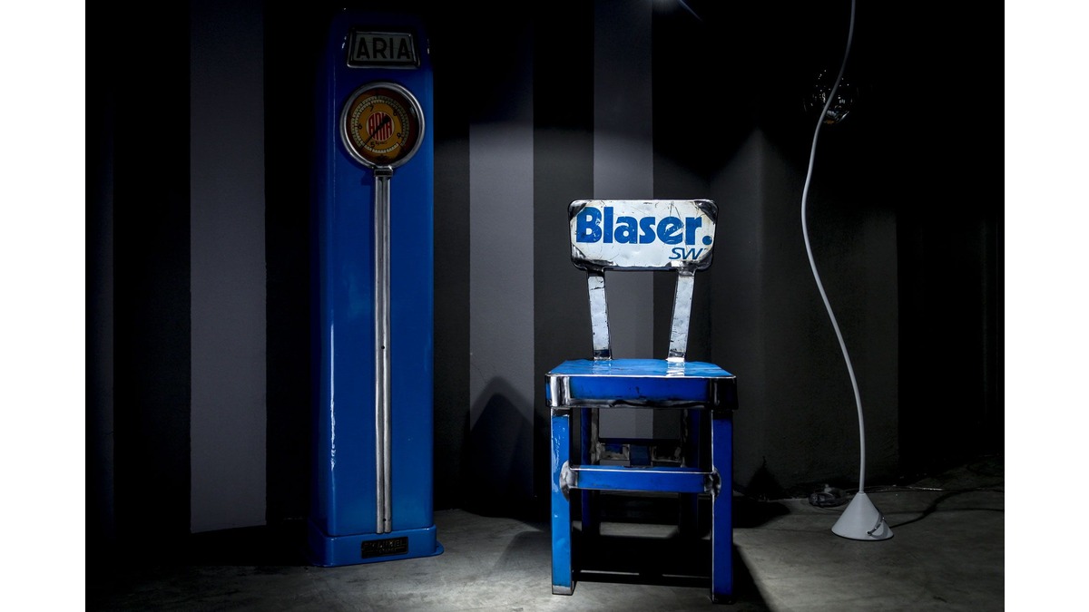 Sedia dora in lamiera di bidone blu con scritta Blaser in una stanza poco illuminata, affiancata da una pompa di benzina aria vintage e una lampada da terra.