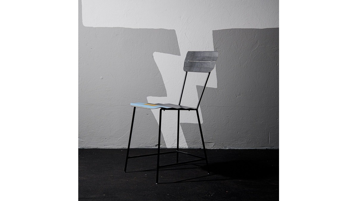 Sedia Wave Chair in lamiera di bidone con seduta blu e schienale grigio in stanza con pavimento nero e muro in cemento grigio.