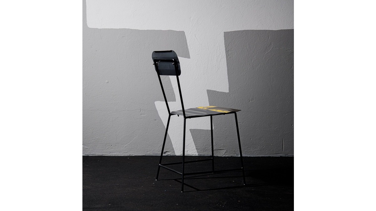 Vista posteriore della Sedia Wave Chair in lamiera di bidone in una stanza con pavimento nero e muro di cemento grigio.