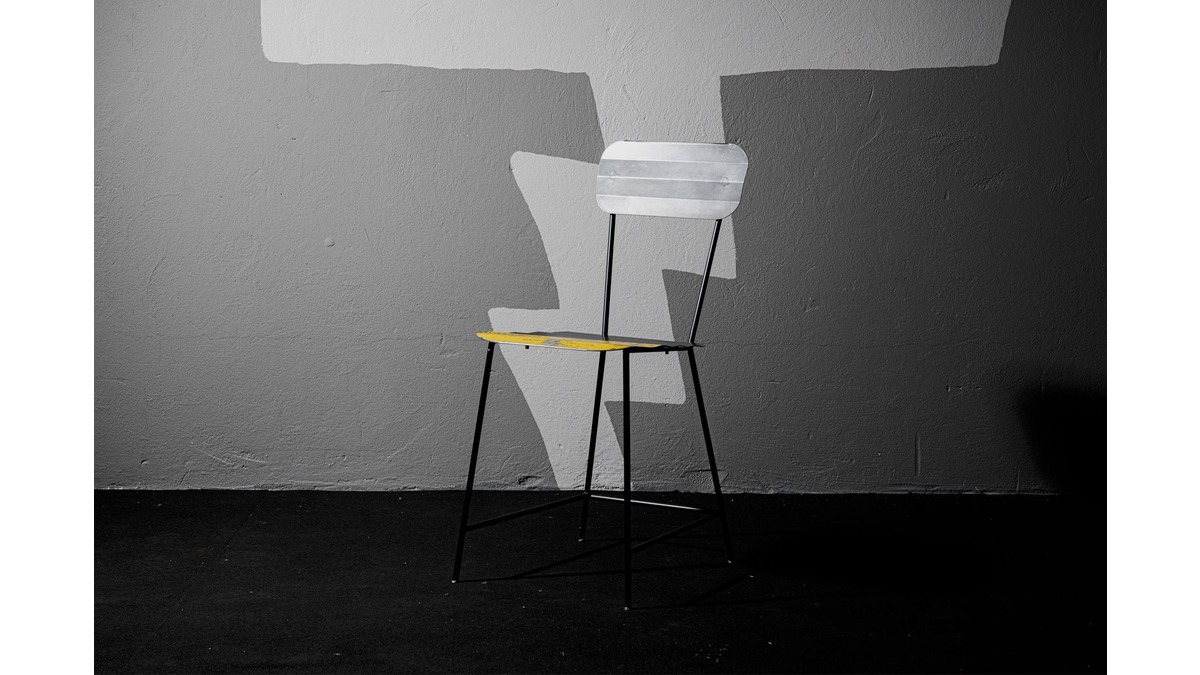Sedia Wave Chair in lamiera di bidone con seduta gialla e schienale grigio in una stanza con pavimento nero e muro in cemento grigio.