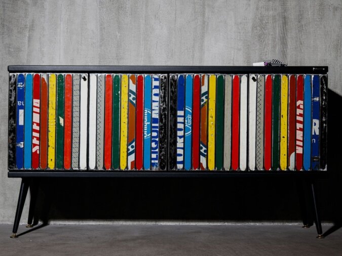 Madia da terra Yoko in lamiera di bidone multicolore con struttura nera in metallo. La madia è situata in uno spazio poco illuminato con muro in cemento grigio.