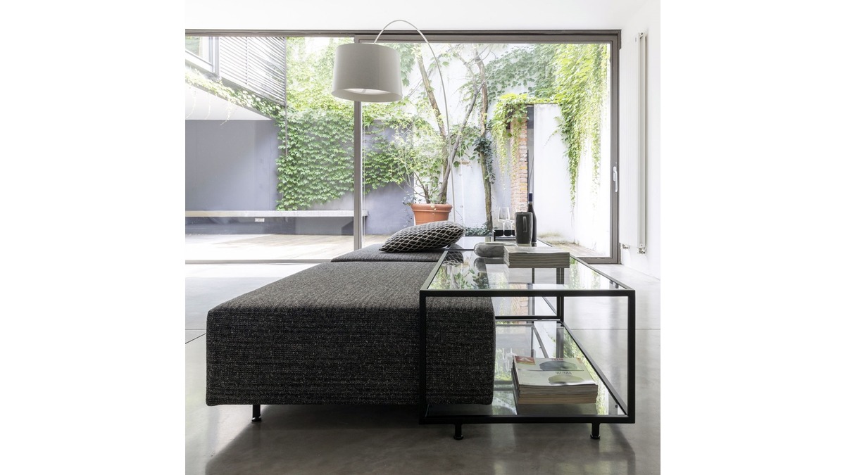 Zoom in living room con pouf imbottito di colore grigio, tavolino in vetro e una grande lampada da terra, che si affaccia su un patio esterno con vegetazione lussureggiante.